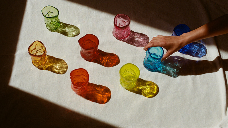 A hand reaches for one of eight, colourful glasses sitting on a linen table cloth in the sunlight
